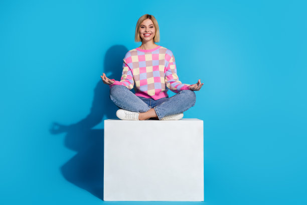 平静好心情女孩的全身照片，穿着彩色套头衫，坐在平台上冥想，在蓝色的背景上孤立的姿势图片下载