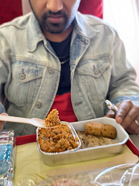 特写照片中，无法辨认的男子坐在飞机座位上，折叠的桌子上放着塑料托盘，里面放着鸡肉串香肠、炒饭和印度烤饼(传统的印度大饼)、塑料瓶水、木叉餐具，重点放在前景上图片下载