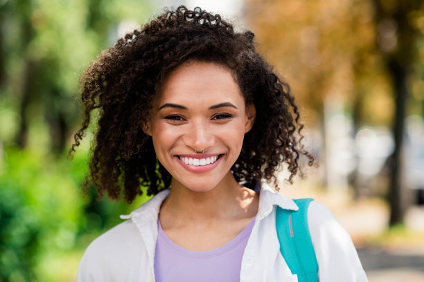 照片中，心情愉快的女士穿着白色衬衫，背着帆布背包，享受阳光明媚的天气，在城市公园外旅行图片下载
