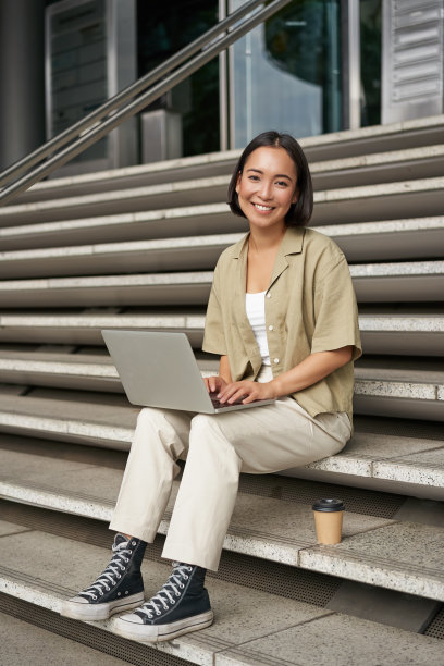 亚洲女孩拿着笔记本电脑坐在大学楼梯上喝咖啡的垂直镜头。年轻女子，学生在户外做作业图片下载