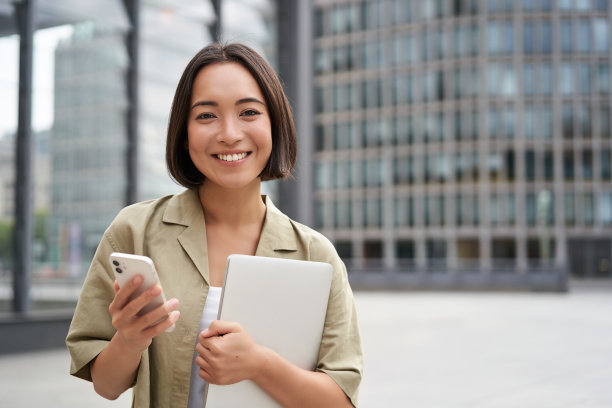 微笑的亚洲女孩带着笔记本电脑，拿着手机，站在街上，看起来很开心图片下载