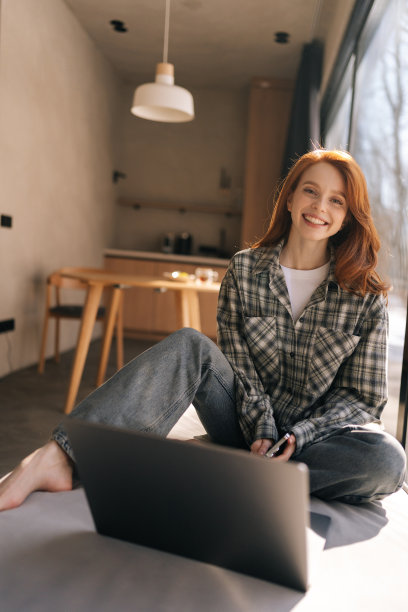 阳光明媚的日子里，快乐的女性自由职业者坐在窗前，用笔记本电脑做研究项目。积极的女远程学生看着相机图片下载