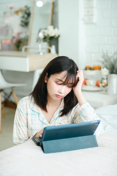 照片中的年轻悲伤的亚洲女性坐在卧室的床边使用数码平板电脑。图片下载