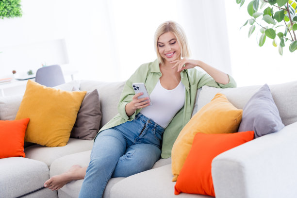 照片中，穿着卡其色衬衫的微笑年轻女子坐在沙发上，用智能手机在家中客厅浏览新闻动态内容信息图片下载