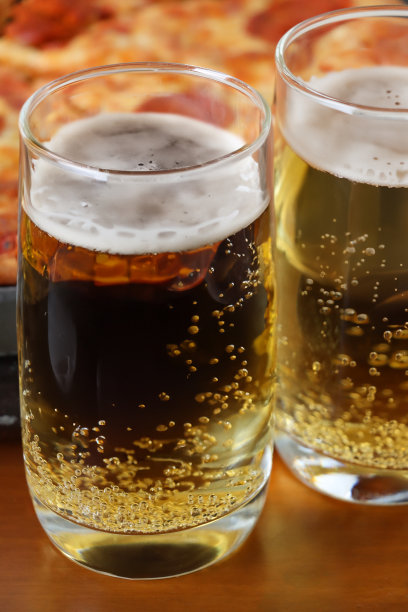餐厅木桌上的两杯半品脱啤酒的特写照片，起泡的气泡形成了泡沫，脆皮切片的意大利辣香肠披萨背景，聚焦在前景上图片下载