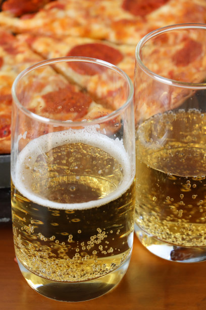 餐厅木桌上两杯半满的半品脱啤酒的特写照片，起泡的气泡形成了泡沫，脆皮切片的意大利辣香肠披萨背景，聚焦在前景上图片下载