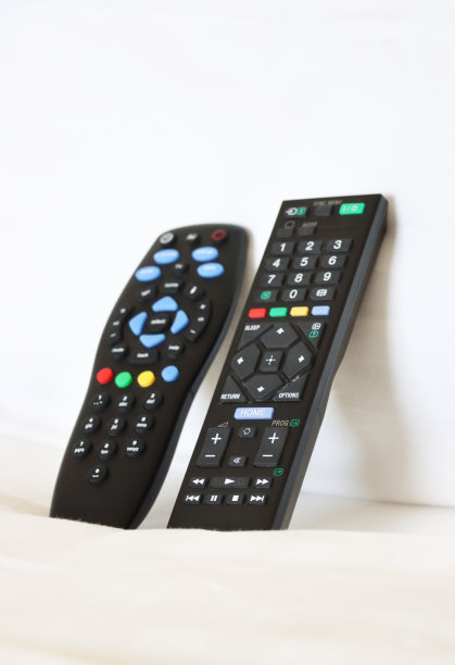 特写图片:两个电视遥控器靠在白色羽绒被上的枕头上，键盘上有红色、绿色、黄色和蓝色的按钮，白色的床上用品背景，重点放在前景上图片下载