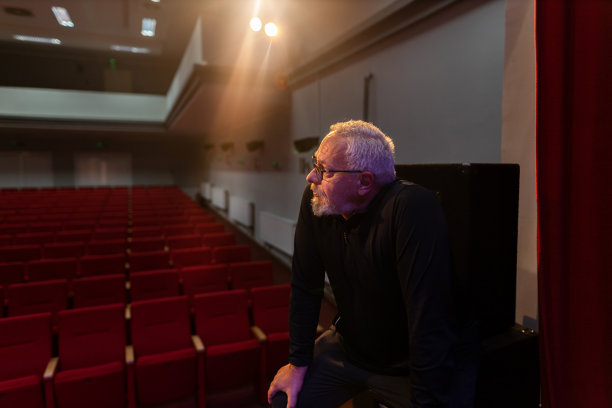 白发苍苍的高级导演，穿着蓝色西装坐在空荡荡的剧院里。红色的椅子。大的对比。看着现场，眼里带着悲伤。库存图片图片下载