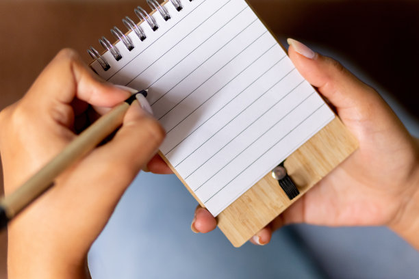 一个女人在笔记本上写东西。这张照片是从上方特写拍摄的，她穿着牛仔裤坐着。图片下载