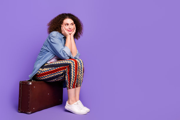 酷漂亮女孩的全尺寸照片，坐在行李箱里，看起来空无一物，穿着牛仔衬衫，孤立在紫罗兰色的背景上图片下载