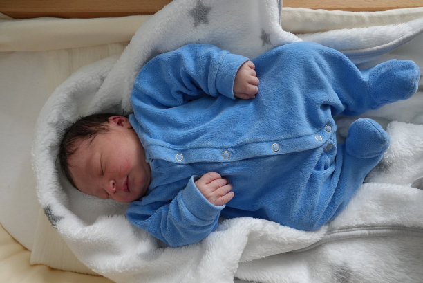 这是一个新生婴儿的全身照片，他安静地睡在婴儿床上，裹着柔软的白色毯子，上面有灰色的星星，穿着蓝色连体衣，体现了宁静和天真。图片下载