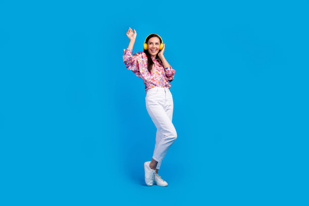 时髦的好心情女士穿着印花衬衫有乐趣享受歌曲耳机孤立的蓝色背景的全长照片图片下载