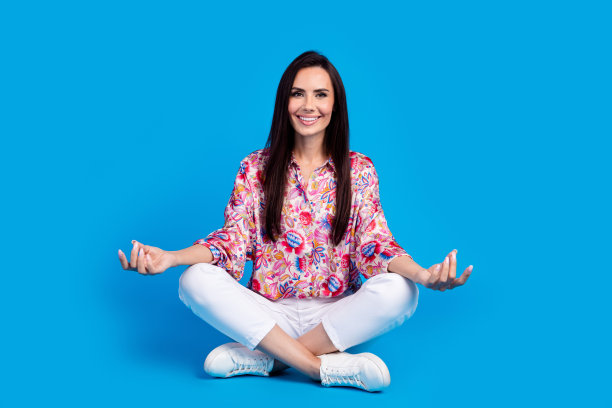 可爱的开朗的女士穿着印花衬衫，双腿交叉，享受瑜伽孤立的蓝色背景的全身照片图片下载