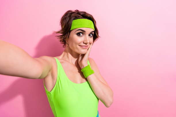 可爱的年轻女士抚摸脸颊自拍照片穿着时尚的绿色运动连衣裤孤立在粉红色的背景图片下载