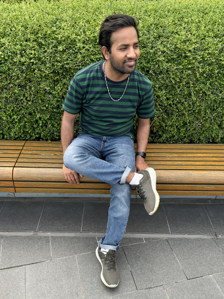 特写照片:印度男子穿着休闲装条纹t恤和牛仔裤，坐在现代木凳上，一条腿交叉在另一条腿上，前面是修剪整齐的树篱，公共铺砌区域，重点放在前景上图片下载