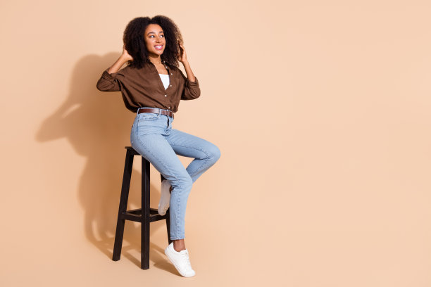可爱女孩的全身照片，穿着棕色衬衫，坐在凳子上，看着提供空白的空间，触摸头发孤立在柔和的颜色背景图片下载