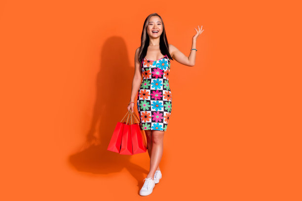 全尺寸照片的狂喜可爱的女人穿着印花时尚的连衣裙拿着购物袋举起手孤立的橙色背景图片下载