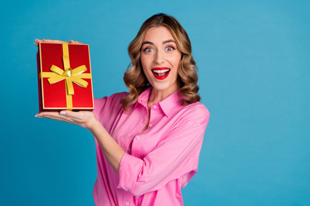 漂亮的年轻女孩的照片肖像拿着兴奋的礼盒，穿着时髦的粉红色衣服，发型孤立在蓝色背景上图片下载