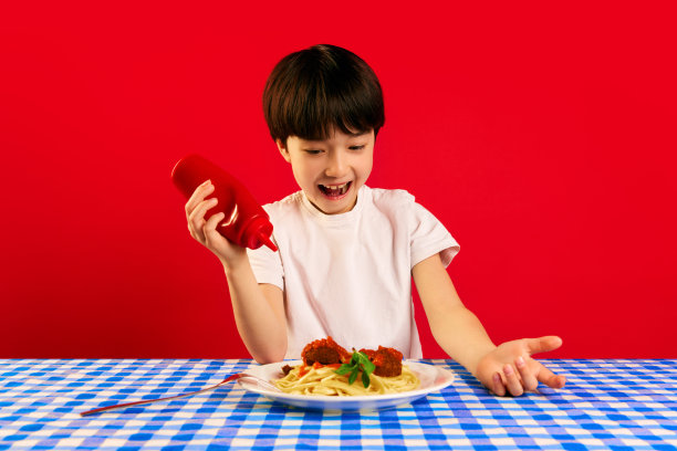 顽皮的孩子，小男孩坐在桌子旁，把番茄酱挤在盘子里，意大利面和肉丸在红色的背景下图片下载