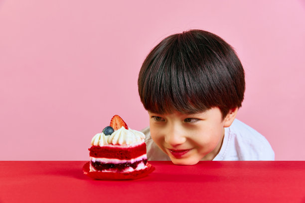 快乐的韩国小男孩把头靠在桌子上，看着粉红色背景下美味的蛋糕图片下载