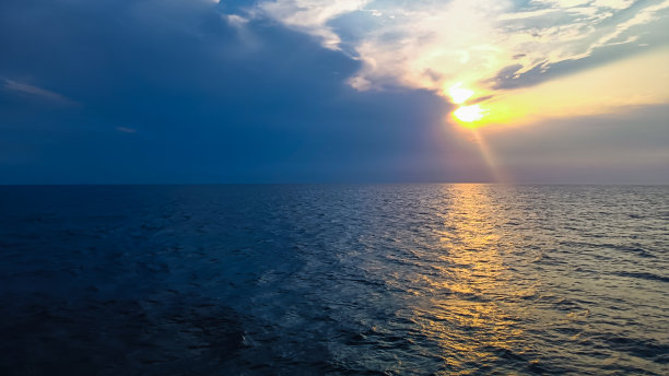 日出或日落时，阳光透过云层照射进来的蓝色海洋。辽阔的大海中平静的水面反射着金色的阳光。象征希望和平静的照片。图片下载