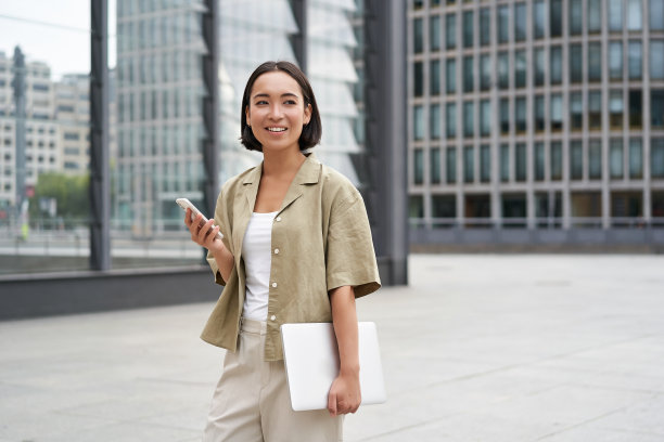 亚洲女孩拿着笔记本电脑和智能手机，站在市中心的街道上，对着镜头微笑图片下载