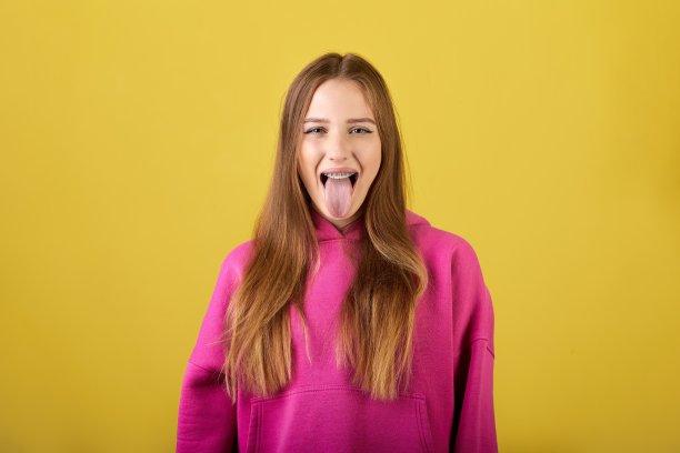 俏皮的年轻女子伸出舌头，穿着黄色背景的粉色连帽衫图片下载