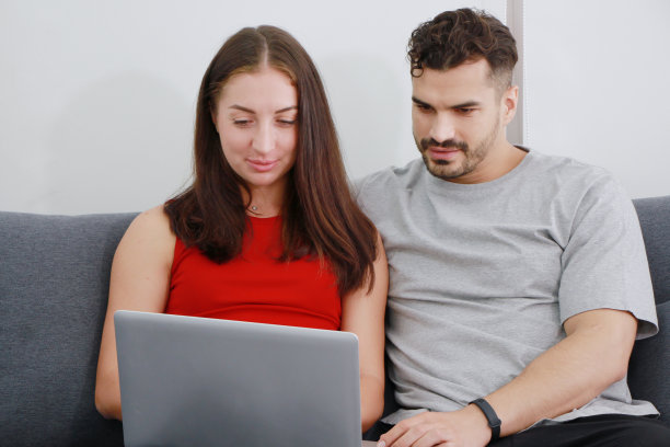 微笑的高加索夫妇喜欢在客厅的沙发上用笔记本电脑在社交媒体上购物。科技与家庭度假生活理念图片下载