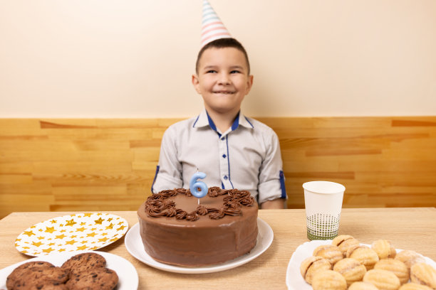 一个男孩坐在桌子前，桌上放着一个生日蛋糕，蛋糕里有一支蜡烛，形状像数字6。生日庆祝的概念。把注意力集中在前面的物体上。图片下载