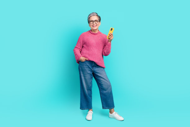 全尺寸照片的聪明的老年人穿着针织套头牛仔裤拿智能手机手臂在口袋里孤立的绿松石色背景图片下载