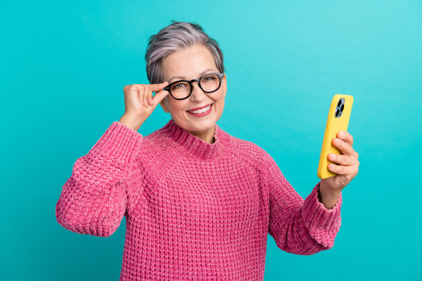 照片中快乐的人穿着针织衫，戴着眼镜，看智能手机，看新闻，手臂放在口袋里，孤立的蓝绿色背景图片下载