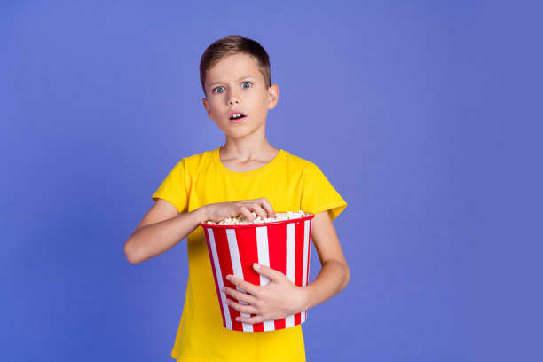 震惊的恐怖男孩的照片，穿着时髦的黄色衣服，拿着篮子美味的爆米花，在紫色的背景下看恐怖电影图片下载