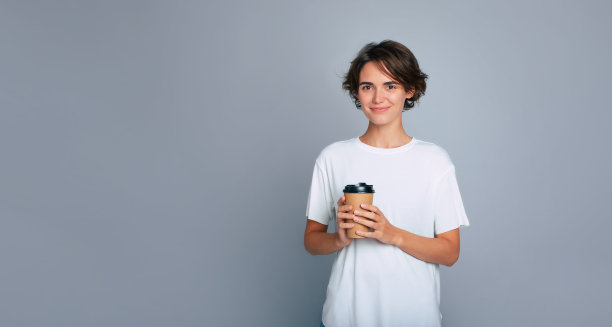美丽的现代女性的特写照片，时髦的发型，白色t恤，微笑着拿着纸杯外卖咖啡，孤立在灰色的背景下。图片下载