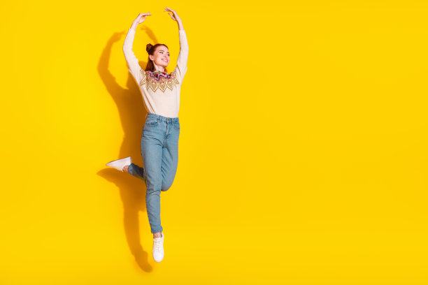 照片肖像漂亮的年轻女子跳芭蕾，举起手穿着时尚的针织温暖的衣服孤立的黄色背景图片下载