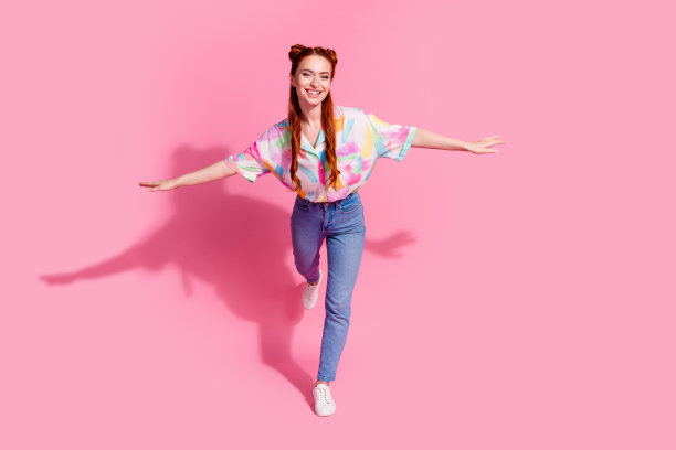 好心情的全尺寸照片漂亮的女孩穿着彩色衬衫牛仔裤牵着手像翅膀在一条腿上孤立的粉红色背景图片下载