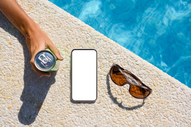 模板假日广告。白色显示屏的手机和在酒店泳池附近喝饮料的度假者的手。有选择性的重点。图片下载