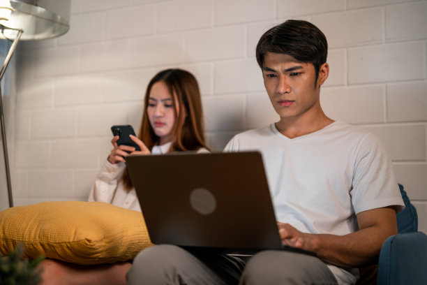 亚洲年轻人晚上和妻子在家里用手机工作。迷人美丽的妻子穿着睡衣等着丈夫用笔记本电脑工作，在家里睡觉时上网交流。图片下载