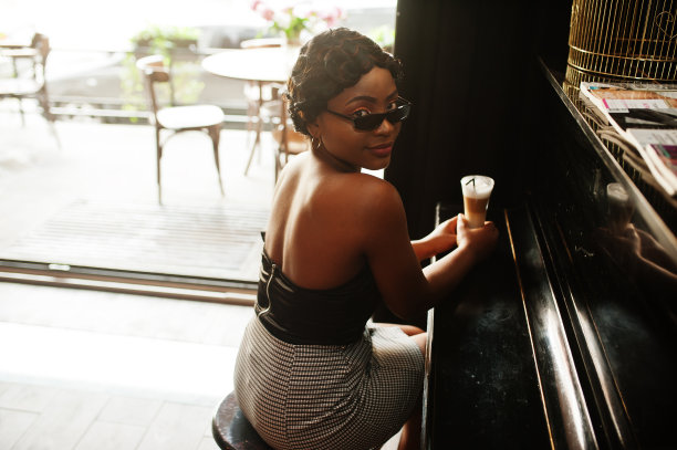 非裔美国女性的肖像，复古发型，拿着一杯拿铁在餐厅摆姿势，坐在钢琴前。图片下载