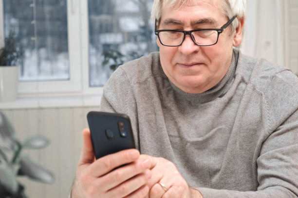 白发苍苍的老人看着网络摄像头，拿着手机，和孩子聊天，老婆上网。老爷爷微笑着与朋友、家人视频聊天。现代科技和老一辈。图片下载