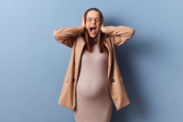 震惊、愤怒、压力大的孕妇穿着漂亮的裙子和夹克，在蓝色背景下独自拍照，捂着耳朵，双手大声尖叫，患有精神疾病图片下载