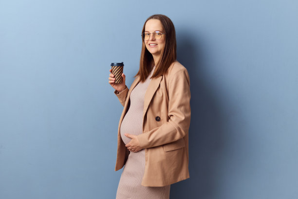 微笑快乐的孕妇，穿着漂亮的裙子和夹克，在蓝色背景下独自摆姿势，享受热外卖咖啡，看着镜头喝着热饮图片下载
