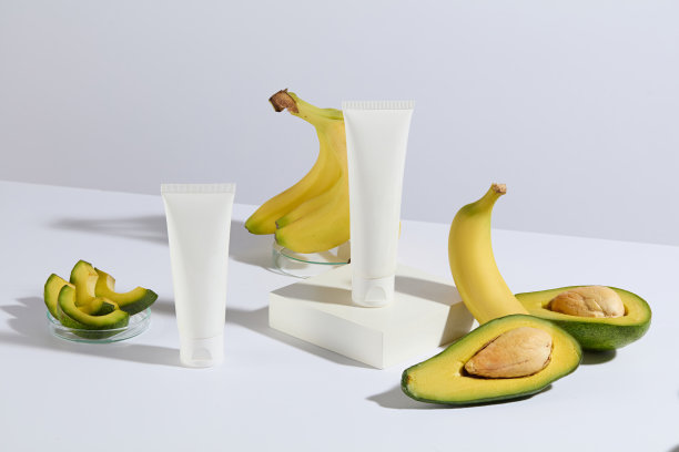 白色色调的牛油果和香蕉组成的照片，两根没有标签的化妆管陈列在桌子上，其中一根放在一个白色的矩形平台上，周围摆放着新鲜的水果图片下载