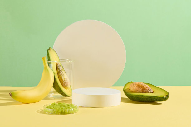 绿色背景，白色圆形背景装饰的宣传照片，香蕉倚在装有半个鳄梨的烧杯上。旁边是一个空白的平台和玻璃盘，上面放着切碎的鳄梨和香蕉图片下载