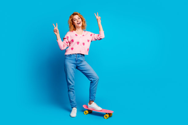 迷人的年轻女子全身照片骑滑板v型标志穿着时尚的粉红色衣服孤立的蓝色背景图片下载