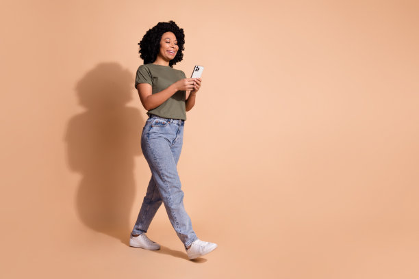 乐观的女性全身照片，穿着卡其色t恤牛仔裤，带着智能手机走在米色背景上，走在空旷的地方图片下载