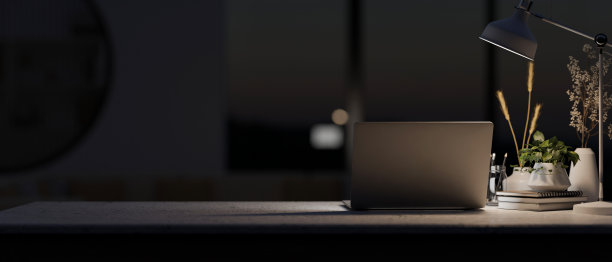 在一个现代的黑暗办公室里，晚上有一台笔记本电脑和桌子上一盏灯的昏暗灯光。图片下载