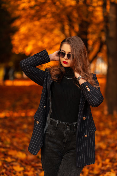 漂亮的女孩戴着时髦的太阳镜，穿着时髦的黑色西装，西装外套和黑色毛衣，走在秋天的公园里，秋天的树叶五颜六色图片下载