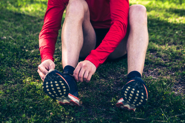 正面照片中，一名身穿红衬衫和黑色短裤的男子正坐在草地上系鞋带图片下载