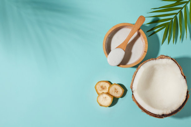 浅蓝色背景照片，高角度拍摄，一个勺子与白色奶油放置在一个木碗，旁边的半个椰子，一些香蕉片和椰子叶装饰。文本复制空间图片下载