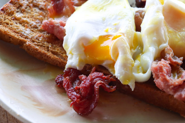 全画幅图片的白色盘子鸡蛋本尼迪克特食谱，一片全麦吐司顶部的培根片，荷包蛋和荷兰酱，渗出的黄色蛋黄，重点在前景图片下载